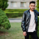 Stylish indian beard man at black leather jacket. India model posed at green garden of city.